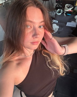 Woman with green eyes, wavy brown-blonde hair, one-shoulder brown top, necklace, Apple Watch, selfie in cluttered room.