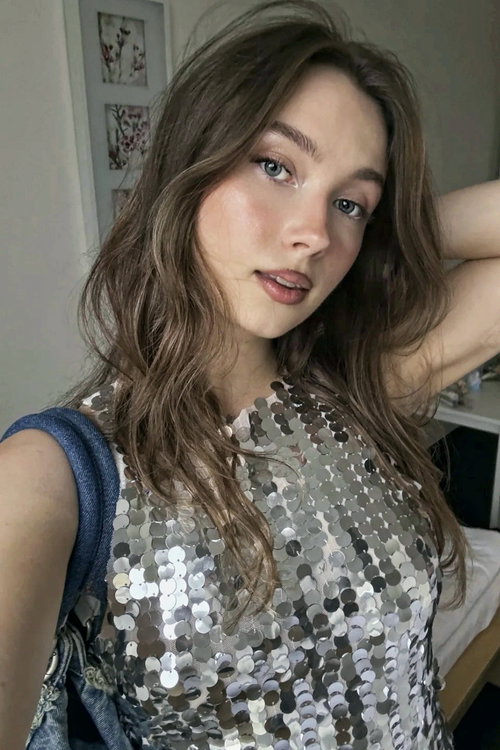 A young woman with wavy brown hair and blue eyes wears a silver sequined top, posing in a bedroom with floral artwork