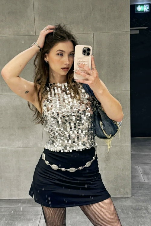A woman with long brown hair takes a mirror selfie in a sequined top, black mini skirt, and fishnet tights, holding a