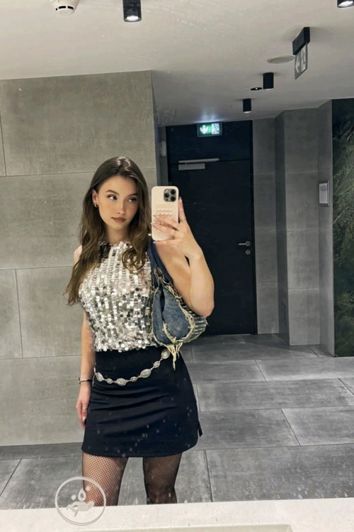 A young woman with long brown hair takes a mirror selfie in a sequined top, black mini skirt, and fishnet stockings in