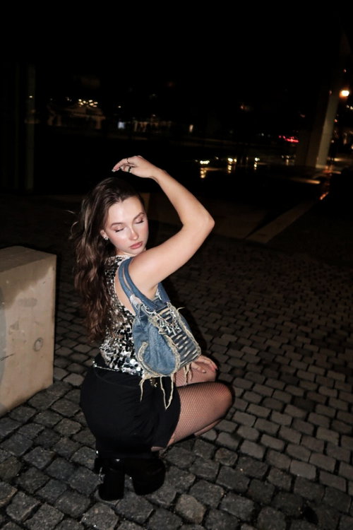 A woman with long brown hair crouches on a cobblestone path at night, wearing a sequined top, denim vest, black skirt,