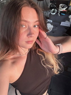 Slim woman with wavy brunette-to-blonde hair in one-shoulder brown top in messy room.