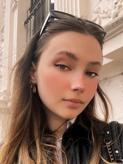 Slim brunette with sunglasses atop head, gold earrings, leather jacket over white shirt against ornate building.