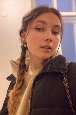 Slim woman with braided brunette hair in hoodie and black jacket, indoor selfie by window.
