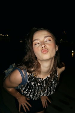 A woman with shoulder-length brown hair poses playfully at night, wearing a sequined top and dark bottoms, hands on