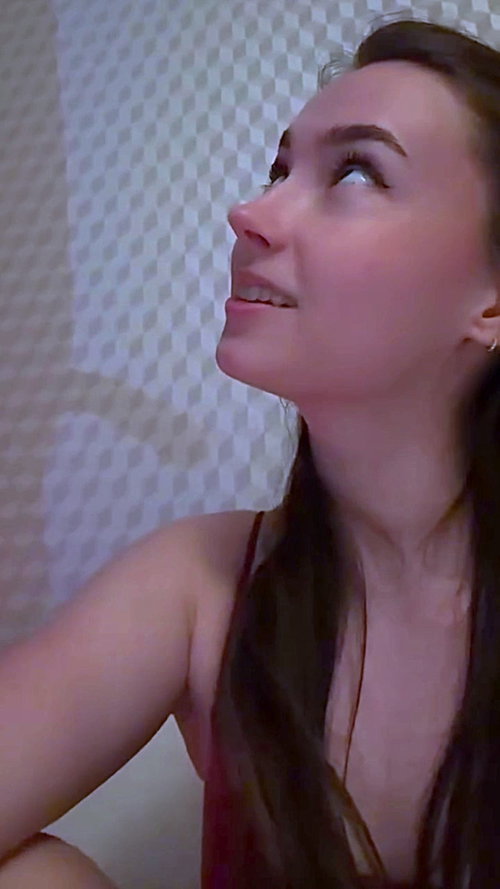 Brunette woman looking upward with parted lips, wearing thin-strapped red top, bare shoulders, tiled bathroom wall.