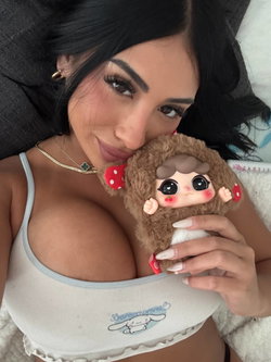 Brunette woman with long dark hair lying down, wearing a white crop top, holding a plush toy close to her chest.