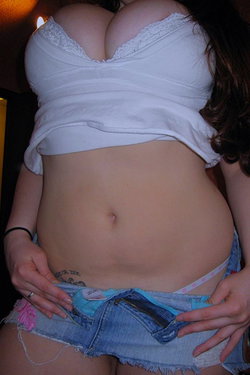 Curvy woman in white lace-trim top and denim shorts, hands on hips, midriff exposed in dimly lit indoor setting.