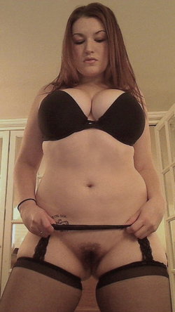 A woman with long brown hair in a black bra and matching panties stands in front of a mirror.