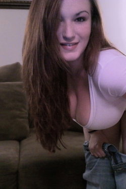 Brunette woman with long hair in a tight white top, holding jeans, posing on a couch indoors.