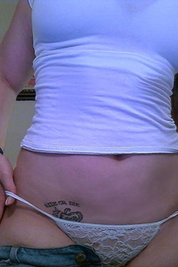 Woman in white tank top and lace thong, hand pulling waistband down, tattoo visible on lower abdomen.