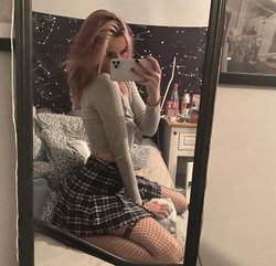 A woman with pink-tinted hair takes a mirror selfie in a beige crop top, plaid skirt, and fishnet stockings, sitting on