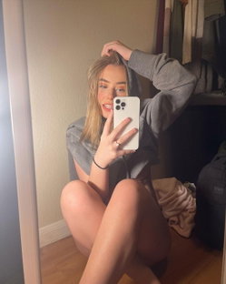 Blonde woman in a gray hoodie takes a mirror selfie, sitting on the floor with legs bent, hand in hair.