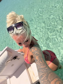 Blonde woman in a pink bikini with full sleeve tattoos poses in clear turquoise water beside a boat, wearing sunglasses