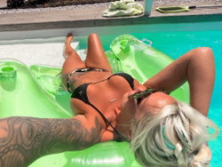 Blonde woman with tattoos lounging on a green float in a pool, wearing a black bikini top and patterned bottoms.