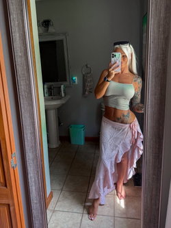 A blonde woman with tattoos takes a mirror selfie in a bathroom, wearing a light green crop top and a pink wrap skirt,