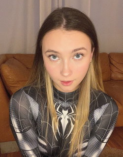 A young woman with long dark hair and light eyes wears a black and white Spider-Man themed bodysuit, sitting on a brown