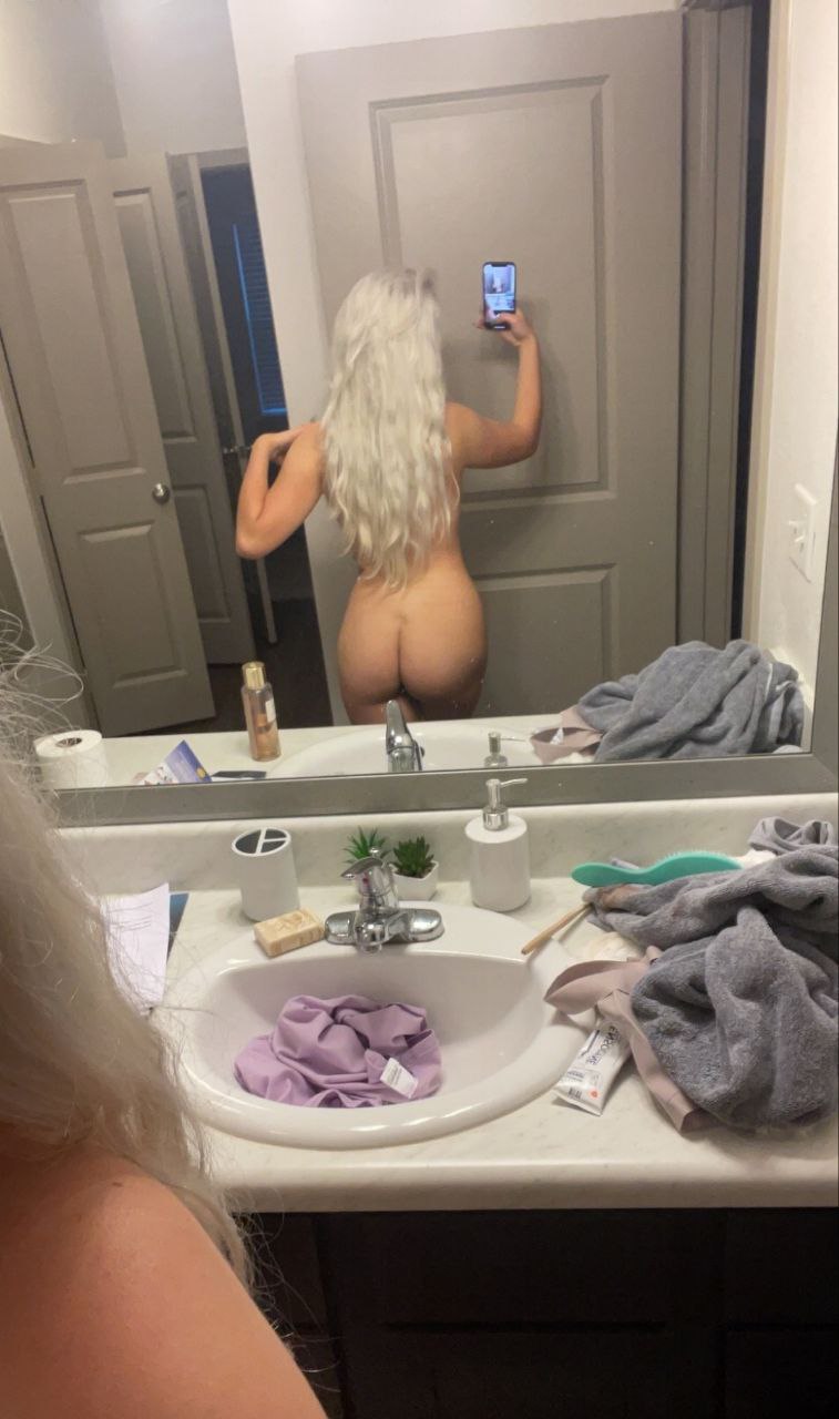 A woman with long platinum hair takes a mirror selfie in a bathroom, showing her bare back and buttocks while holding