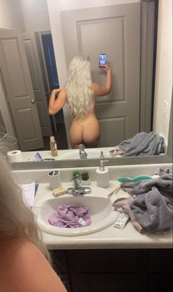 A woman with long platinum hair takes a mirror selfie in a bathroom, showing her bare back and buttocks while holding