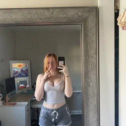 Redheaded woman in a gray tank top and sweat shorts takes a mirror selfie, posing with one hand near her lips.