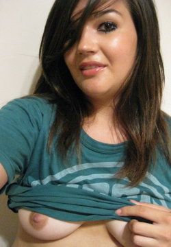 Brunette woman with long hair in a teal t-shirt, lifting the shirt to expose bare breasts, taking a close-up selfie