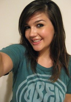 A smiling woman with shoulder-length dark hair wearing a green graphic t-shirt takes a selfie indoors.