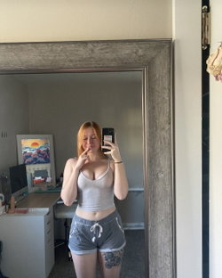 Redheaded woman in a white crop top and gray shorts takes a mirror selfie, showing a floral tattoo on her thigh in a