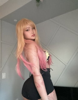 A person with long blonde and pink hair in a black outfit poses in a modern room.